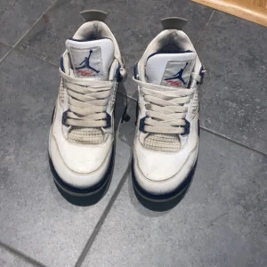 Nike Air Jordan 4 Retro Vit/Blå - Säljer ett par Nike Air Jordan 4 Retro sneakers i vitt med blå detaljer och röd text på plösen. Skorna har klassisk mesh och skinn, snörning och en chunky sula. Perfekta för dig som gillar ikoniska och sportiga sneakers med streetkänsla.