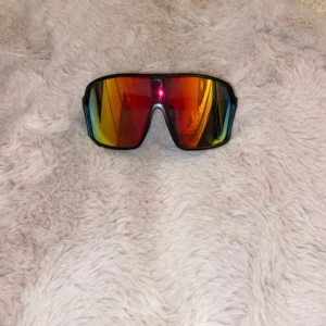 Sportiga solglasögon från Noixx - Säljer ett par sportiga solglasögon från Noixx med spegelglas i rött, gult och blått. Bågarna är svarta och modellen är bred och omslutande, perfekt för utomhusaktiviteter. Linsen har en cool spegeleffekt och ger ett futuristiskt intryck.