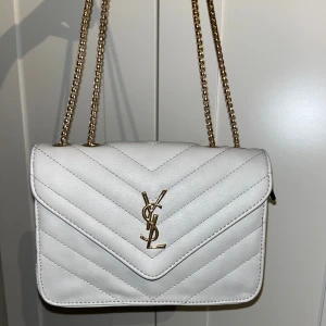 Vit quiltad axelväska från YSL - Snygg vit axelväska från YSL med guldiga kedjor och logga framtill. Väskan är quiltad i ett V-mönster och har ett rymligt vinrött innerfack med dragkedja. Perfekt storlek för det viktigaste och ger en lyxig vibe till din outfit.