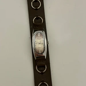 Vintage Regal klocka med brunt läderarmband - Snygg klocka från Regal med avlångt silverfärgat ur och brunt läderarmband. Armbandet har coola metallringar som detaljer och spänne i silver. Urtavlan är enkel med siffror vid 12 och 6, och resten markerat med prickar.