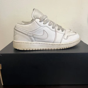 Nike Air Jordan 1 Low vita sneakers - Säljer ett par klassiska Nike Air Jordan 1 Low i helvitt skinn. Skorna har perforerad tå, rund tå, snörning och ikoniska swoosh-loggan på sidorna. Perfekta för dig som gillar en clean och tidlös look. Kommer med originalkartong.