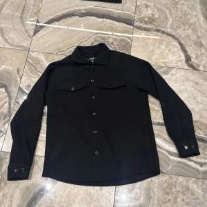 Svart overshirt från SMOG, relaxed fit - Svart overshirt från SMOG i relaxed fit med klassisk krage, två bröstfickor med lock och knappar längs hela framsidan. Långärmad modell med knappar vid ärmslut. Perfekt för en clean och stilren look.