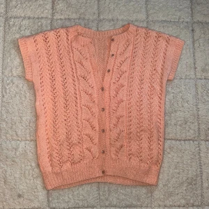 Stickad persikofärgad kofta - Superfin stickad kofta i persikofärg med korta ärmar och snyggt hålmönster. Koftan har rund halsringning och knäpps framtill med små knappar. Perfekt för lager-på-lager och ger en söt vintagekänsla till din outfit.