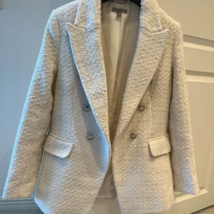 Vit kavaj från H&M i bouclé - Snygg vit kavaj från H&M i bouclé-tyg med dubbelknäppning och klassiska slag. Jackan har två fickor med lock framtill och en strukturerad, lite lyxig känsla. Perfekt för dig som gillar stilrena och trendiga plagg med lite edge.