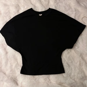 Svart topp med vid passform från Nelly - Svart topp från Nelly i storlek XS med lös och vid passform upptill och smalare midja. T-shirten har rund halsringning och är tillverkad i mjukt material, perfekt för en chill och stilren look.