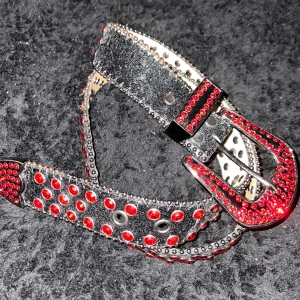 Svart skärp med röda nitar och strass - Coolt svart bälte i skinn med massor av röda strass-stenar och nitar längs hela bandet. 114cm långt med buckle