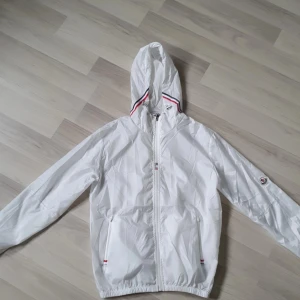 Vit vindjacka från chase med huva - Snygg vit vindjacka från Chase med huva och dragkedja framtill. Jackan har Moncler-logga på ärmen och detaljer i rött, vitt och marinblått vid huvan och fickorna. Tillverkad i lätt polyester, perfekt för blåsiga dagar. Sportig och stilren look.