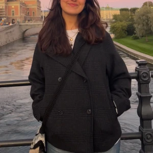 Svart rutig skepparkavaj i ull - Säljer en svart skepparkavaj med diskret rutigt mönster och dubbelknäppning. Jackan har bred krage, långa ärmar och en loose passform som ger en chill vibe. Materialet är mjuk ull och perfekt för höst och vinter. Snygg att styla med jeans och boots.