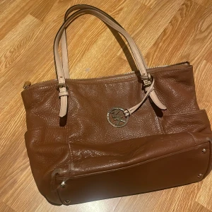 Brun handväska från Michael Kors - Snygg handväska i brunt skinn från Michael Kors med ljusa handtag och metallfästen. Väskan har MK-logga i silver och praktisk dragkedja upptill. Perfekt storlek för att få plats med allt du behöver och stilren design som lyfter din look. Skick se på bilderna sliten på vissa ställen men inget störande men äkta