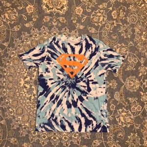 Tie-dye Superman t-shirt från H&M - Cool t-shirt från H&M med blått och vitt tie-dye mönster och stor orange Superman-logga på bröstet. Klassisk rund hals och korta ärmar. Perfekt för dig som vill sticka ut med en färgstark och unik look.