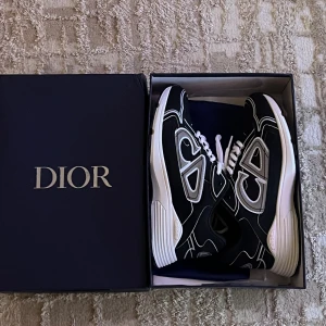 Svarta Dior B30 sneakers strl 42 - Säljer ett par svarta Dior B30 sneakers i mesh och tekniskt tyg. Skorna har vita snören, vita och grå detaljer samt en chunky vit sula. Stora Dior-loggor på sidorna och coolt mönster i kontrastfärger. Perfekt för dig som gillar exklusiva och trendiga sneakers.