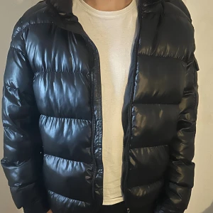 Svart Moncler pufferjacka med huva - Svart pufferjacka från Moncler med glansig finish, huva och dragkedja framtill. Jackan har en ficka med Moncler-logga på ärmen. Huvan har ett litet hål men syns inte och jag kan även sy ihop det innan säljning ifall du vill det. Dragkedjan är även sönder så jackan går ej att stänga. Skriv privat för fler bilder om äkthet.