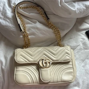 Beige quiltad Gucci axelväska - Säljer en beige Gucci Marmont axelväska i quiltat skinn med guldiga kedjor och GG-logga framtill. Väskan har ett hjärtdetalj på baksidan och röd insida med dragkedja. Perfekt storlek för det viktigaste och riktigt snygg design.