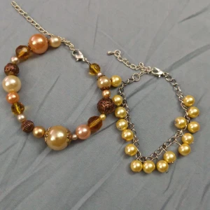 Två armband med pärlor i guld och brunt - Säljer två snygga armband med pärlor i olika storlekar och färger. Ett armband har mixade pärlor i guld, brunt och champagne på en silverkedja, medan det andra har guldfärgade pärlor hängande på en länkad kedja. Perfekt för dig som gillar unika accessoarer! Båda armbanden har förlängningskedja och passar jättebra att bära tillsammans.
