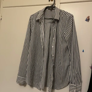 Randig vit och svart skjorta - Snygg skjorta med vertikala svarta och vita ränder, klassisk krage och knappar framtill. Långärmad med breda manschetter och knappar även vid ärmslut. Skjortan har en loose passform och är tillverkad i mjuk bomull som känns skön mot huden.