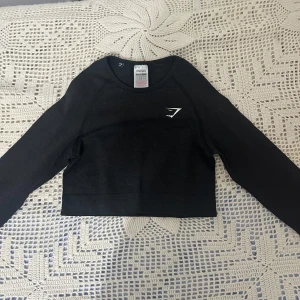 Svart croppad långärmad topp Gymshark - Svart croppad långärmad topp från Gymshark med diskret logga på bröstet. Tight passform och rund halsringning, perfekt för träning eller chill. Tillverkad i mjukt och stretchigt material som ger bra rörelsefrihet.