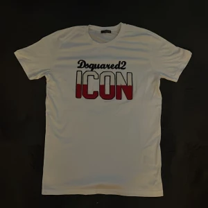 Vit Dsquared2 ICON t-shirt i bomull - Vit t-shirt från Dsquared2 med stor, broderad 'ICON'-text i vitt och rött framtill. Klassisk passform med korta ärmar och rund hals. Tillverkad i 100% bomull för en skön känsla. Snygg kontrast mellan svart, vitt och rött i trycket. Saknar tag, helt ny aldrig använd