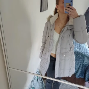Beige pufferjacka med fuskpäls - Säljer en beige pufferjacka med fuskpäls på kragen och snygga detaljer som bälte i midjan och stora fickor med knappar. Har knapt använt den. Men den har några små fäkar som knapt syns. Perfekt för kalla dagar och har en riktigt chill vibe.