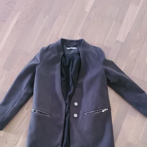 Svart kavaj från Gina Tricot, stl 34 - Stilren grå kavaj från Gina Tricot med lång ärm, två dekorativa knappar framtill och dragkedjedetaljer vid fickorna. Kavajen har en rak passform och är fodrad med svart tyg. Perfekt för en snygg och clean look.