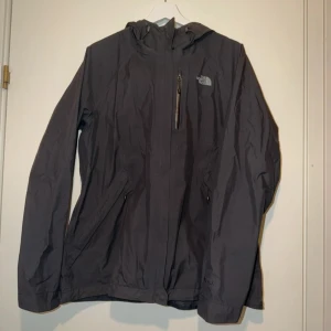 Svart vindjacka från The North Face nyskick - Svart vindjacka från The North Face med huva och dragkedja framtill. Jackan har tre fickor med dragkedja och diskret logga på bröstet. Tillverkad i polyester och nylon, perfekt för blåsiga dagar. Snygg och funktionell design. Storlek xl