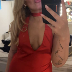 Röd långklänning med djup urringning - Säljer en röd långklänning i satinliknande material med djup v-ringning och öppen rygg. Klänningen har en snygg knyt-detalj i nacken och slits nertill. Perfekt för dig som vill sticka ut med en elegant och trendig look.