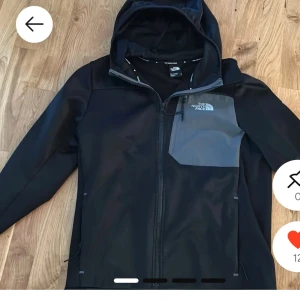 Svart vindjacka från The North Face - Svart vindjacka från The North Face med grått bröstparti och logga. Jackan har huva, hel dragkedja och praktisk bröstficka med dragkedja. Materialet är syntet och insidan är fodrad med mjuk fleece. Perfekt för blåsiga dagar.