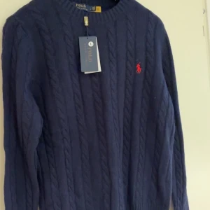 Marinblå kabelstickad tröja Polo Ralph Lauren - Kabelstickad marinblå tröja från Polo Ralph Lauren med klassisk röd broderad logga på bröstet. Tröjan har rund halsringning och långa ärmar. Perfekt för dig som gillar en tidlös och stilren look.