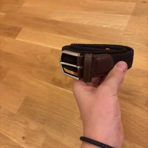 Flätat bälte från John Henric - Snyggt flätat bälte från John Henric i mörkbrunt och marinblått med detaljer i skinn. Bältet har en stilren silverfärgad metallspänne och är markerat med 39/100. Ser inte ut som dehär vanliga temu bältet vilket skapar lite karaktär. 