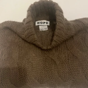 Brun polotröja i ull från HOPE - Säljer en chunky brun polotröja från HOPE med grov kabelstickning. Tröjan har hög krage, lång ärm och är stickad i mjuk ullblandning. Perfekt för kalla dagar och riktigt snygg till jeans eller kjol.