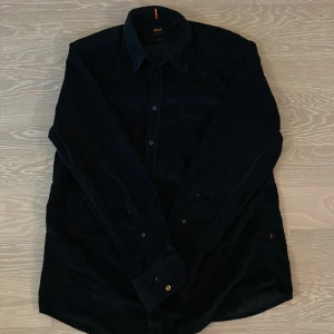 Svart manchesterskjorta från BOSS - Svart skjorta från BOSS i mjuk manchester med klassisk krage och knappstängning framtill. Regular fit och långärmad modell med bröstficka där BOSS-loggan är diskret broderad. Perfekt för dig som gillar stilrena och bekväma plagg.