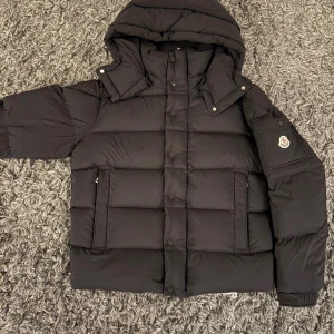 Svart Moncler pufferjacka med huva - Svart pufferjacka från Moncler med huva, stora fickor och ikonisk logga på ärmen. Jackan har både dragkedja och knappar framtill, samt quiltad design som ger en schysst volym. Perfekt för kalla dagar och har en clean, modern look.