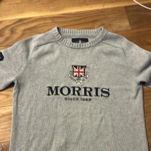Grå stickad tröja från Morris S - Snygg grå stickad tröja från Morris med broderad brittisk flagga och text på bröstet. Tröjan har ribbade muddar, rund hals och svart 68-tryck på ena ärmen. Perfekt för dig som gillar klassisk och sportig stil. Har dock två små defekter på loggan men de syns inte om man inte nämner det