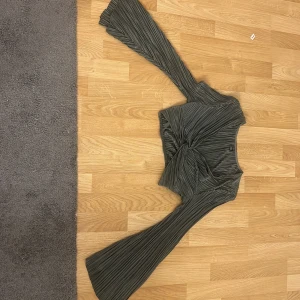 Grå plisserad croppad topp Shein - Säljer en grå plisserad croppad topp från Shein i storlek XS. Toppen har långa utsvängda ärmar och en snygg twist-detalj framtill. Materialet är glansigt och lätt, vilket ger en cool och trendig look.