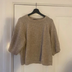 Beige stickad tröja med halvärm - Mysig beige stickad tröja med rund hals och halvärmar. Tröjan har en loose passform och är perfekt för lager-på-lager under kyliga dagar. Materialet är mjukt och ger en chill vibe till din outfit.