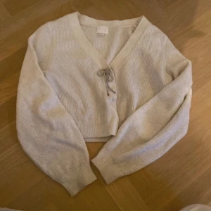 Beige croppad kofta med knyt - Superfin beige croppad kofta med ribbad struktur och v-ringad hals. Tröjan har långa ärmar och en söt knytning framtill. Perfekt att slänga över en topp för en chill look. Passar dig som gillar mjuka och neutrala färger.