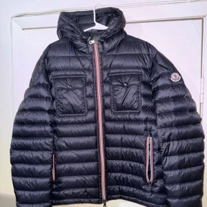 Svart dunjacka från Moncler - Säljer en svart dunjacka från Moncler med huva och två bröstfickor med knappar. Jackan har en snygg rödvit dragkedja framtill och Moncler-logga på ärmen. Perfekt för kalla dagar och har en puffad look som är riktigt trendig.