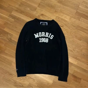 Svart stickad tröja Morris 1968 - Svart stickad tröja från Morris i storlek Large med vit text 'Morris 1968' på bröstet. Tröjan har rund hals och långa ärmar, samt ribbade muddar vid ärmslut och nederkant. Perfekt för dig som gillar klassisk och sportig stil.