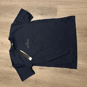 Mörkblå Stone Island t-shirt - Mörkblå t-shirt från Stone Island med logga tryckt på bröstet och en stor, rund logga i silver på ryggen. Klassisk passform med rund hals och korta ärmar. Perfekt för dig som gillar stilrena streetwear-plagg.