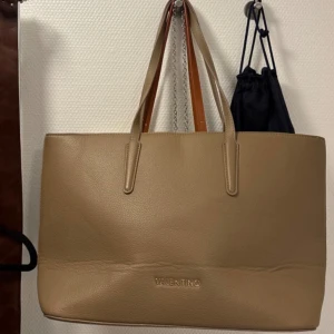 Beige shoppingväska från Valentino - Stilren och rymlig tote bag från Valentino by Mario Valentino i klassisk beige färg. Väskan är i fint skick och passar perfekt både till vardag och jobb. ✨ Elegant design med präglad logotyp framtill ✨ Gott om plats för dator, böcker eller allt du behöver till vardagen ✨ Dragkedja upptill för extra säkerhet ✨ Märkt med äkta etikett och innerfoder från Mario Valentino (Mirade S.p.A.)  Mått ca: 📏 Bredd: 40 cm 📏 Höjd: 28 cm 📏 Djup: 14 cm 📏 Handtag drop: ca 24 cm