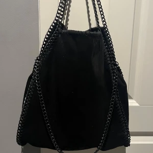 Svart axelväska med kedjedetaljer - Säljer min snygga svart axelväska som jag inte använder längre. Väskan har coola kedjedetaljer längs kanterna och som axelrem. Väskan har en mjuk mockaliknande tyg. 