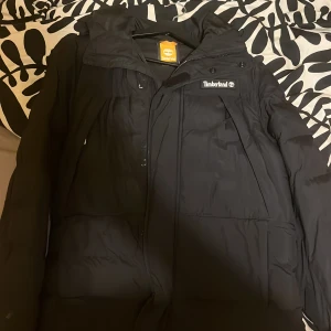 Timberland Vinterjacka (helt ny) - Säljer en svart vinterjacka från Timberland med huva och synlig logga på bröstet. Jackan har dragkedja och knappar framtill, samt flera fickor. Perfekt för kalla dagar och har en stilren look som funkar till det mesta.