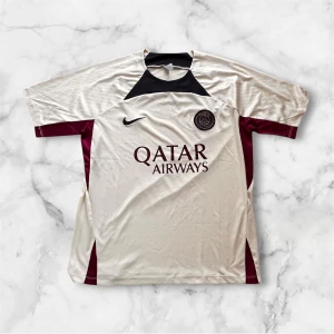 PSG fotbollströja från Nike - Paris Saint-Germain fotbollströja i ljus beige med mörkblå och vinröda detaljer. Tröjan har korta ärmar, broderat PSG-emblem och Nike-logga på bröstet samt Qatar Airways-tryck. Tillverkad i lätt och ventilerande polyester, perfekt för match eller träning. Nypris ca 550kr: Mitt pris 299kr!