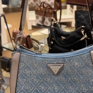 Blå axelväska från Guess med mönster - Snygg blå axelväska från Guess med silverfärgade detaljer och dragkedja. Väskan har ett rutigt mönster med logotyptryck och ljusgrå kanter i skinnimitation. Perfekt storlek för det viktigaste och har en extra ficka med dragkedja på baksidan.