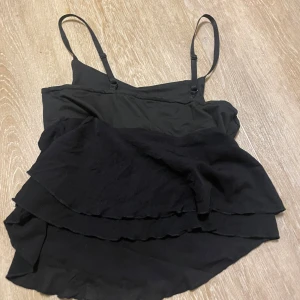 Svart linnetopp med volanger - Säljer en svart linnetopp med smala justerbara axelband och dubbla lager volanger nedtill. Toppen har en mjuk, flowy passform och är perfekt för dig som gillar en enkel men ändå cool look. Materialet känns lätt och skönt mot huden.