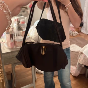 Svart axelväska med gulddetalj - Snygg svart axelväska med mjuk form och guldfärgad metallspänne. Väskan har ett klassiskt, vintage-inspirerat utseende med rejäla axelband och rymligt fack. Perfekt för dig som gillar stilrena accessoarer med en touch av retro.