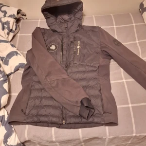 Svart jacka från Sail Racing - Svart jacka från Sail Racing med huva och dragkedja framtill. Jackan har quiltad framsida och rygg, släta ärmar och två sidofickor med dragkedja. Märkeslogga på bröstet och patch på ärmen. Perfekt för kyliga dagar och snygg till streetwear.