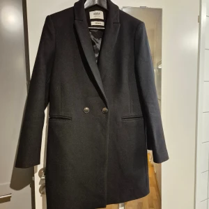 Svart kappa från promod.com, strl 38 - Stilren svart kappa från GISELE med klassisk krage och två snygga knappar framtill. Materialet är vävt i Italien av Manteco och är gjort av ull, vilket ger en exklusiv känsla. Kappan har raka linjer, diskreta fickor och är helfodrad. Perfekt för dig som gillar minimalistisk och elegant stil.