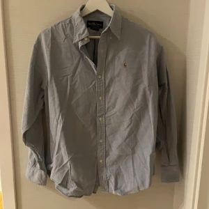 Ljusgrå skjorta från Ralph Lauren - Klassisk ljusgrå skjorta från Ralph Lauren med broderad logga på bröstet. Skjortan har lång ärm, knappar hela vägen och en snygg krage. Tillverkad i mjuk bomull som känns skön mot huden. Perfekt för dig som gillar stilrena och tidlösa plagg. S/XS
