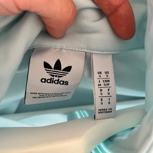 Ljusblå Adidas track kofta  S - Snygg ljusblå Adidas track jacket med klassiska vita ränder längs ärmarna och broderad logga på bröstet. Jackan har hel dragkedja, högt krage och två fickor med dragkedja. Tillverkad i mjuk polyester, perfekt för chill eller sport.