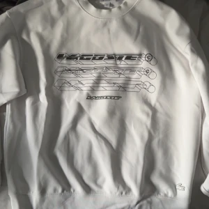 Vit sweatshirt från Lacoste loose fit - Vit sweatshirt från Lacoste i loose fit med coolt grafiskt tryck på bröstet och klassisk rund hals. Tröjan har långa ärmar och den ikoniska krokodilloggan broderad vid nederkanten. Perfekt för dig som gillar stilren streetwear.